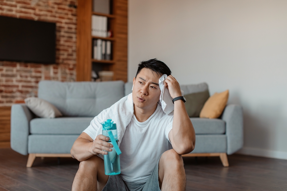 A man sitting on a living room floor holding a water bottle and wiping sweat off his face.